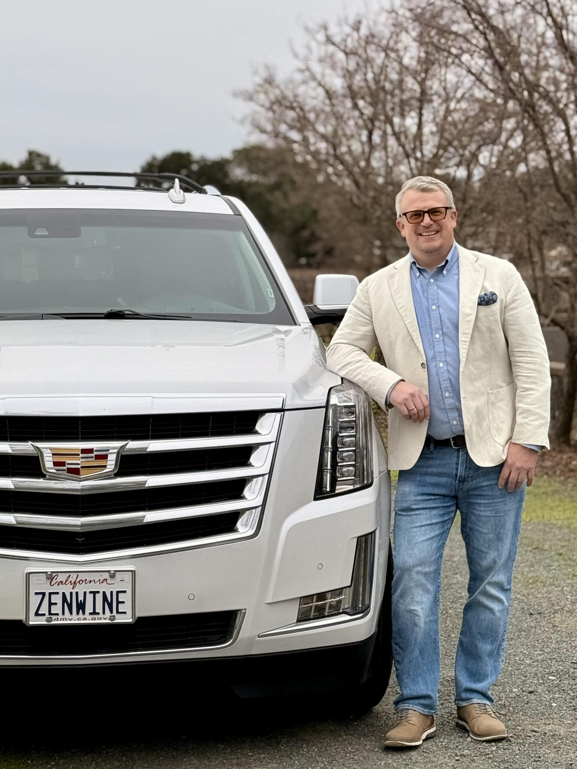 Zen Wine Tours Man in blazer and jeans smiling while standing beside a white SUV with personalized license plate in an outdoor setting.